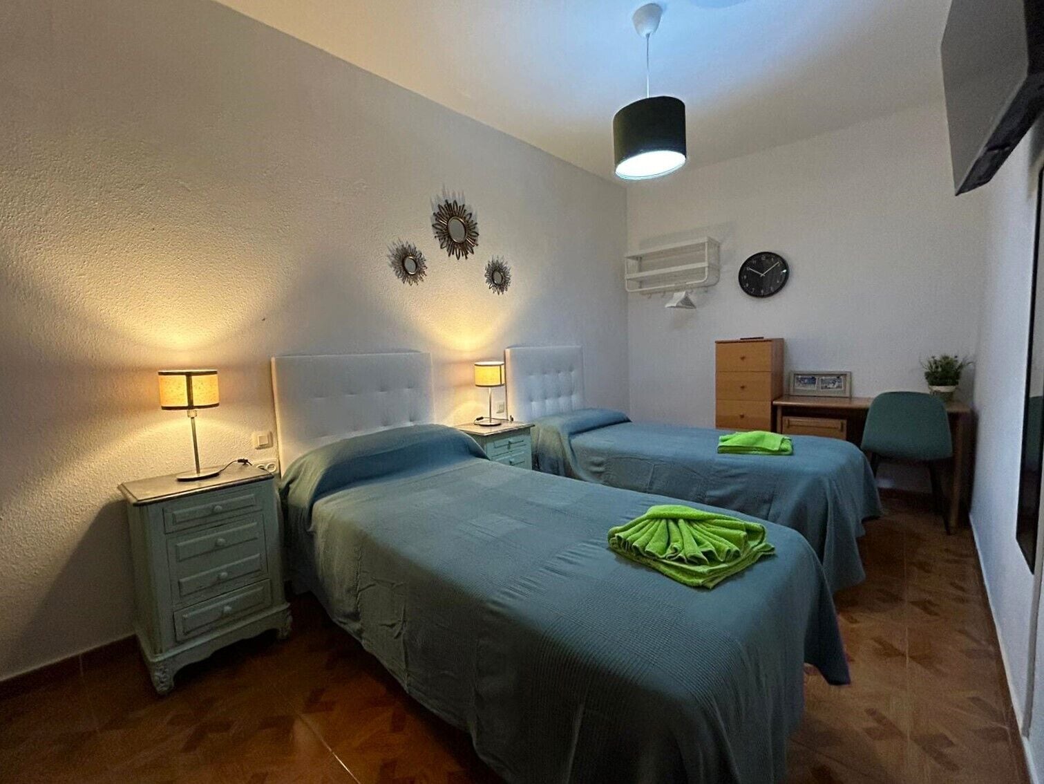 Een slaapkamer met twee bedden, een nachtkastje, een kaptafel, een bureau, een stoel, een klok en wanddecoraties.