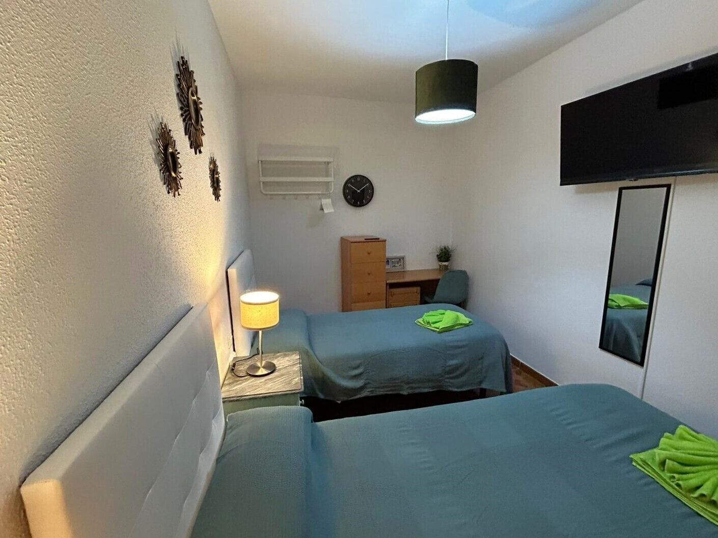 A small, well-lit bedroom with two single beds, a desk, a chair, a TV, and a mirror.