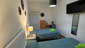 Un dormitorio pequeño y bien iluminado con dos camas individuales, un escritorio, una silla, un televisor y un espejo.