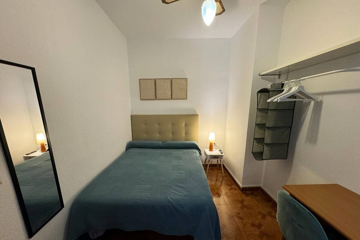 Een ruime, goed verlichte slaapkamer met een bed, nachtlampjes, een spiegel, een dressoir en een kledingrek.