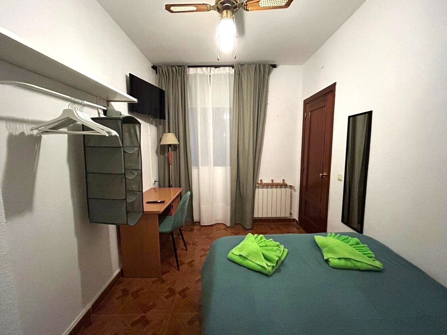 Habitació d'hotel petita i ordenada, amb un llit, una taula de treball, una cadira, un penjador, una làmpada i una finestra amb cortines.