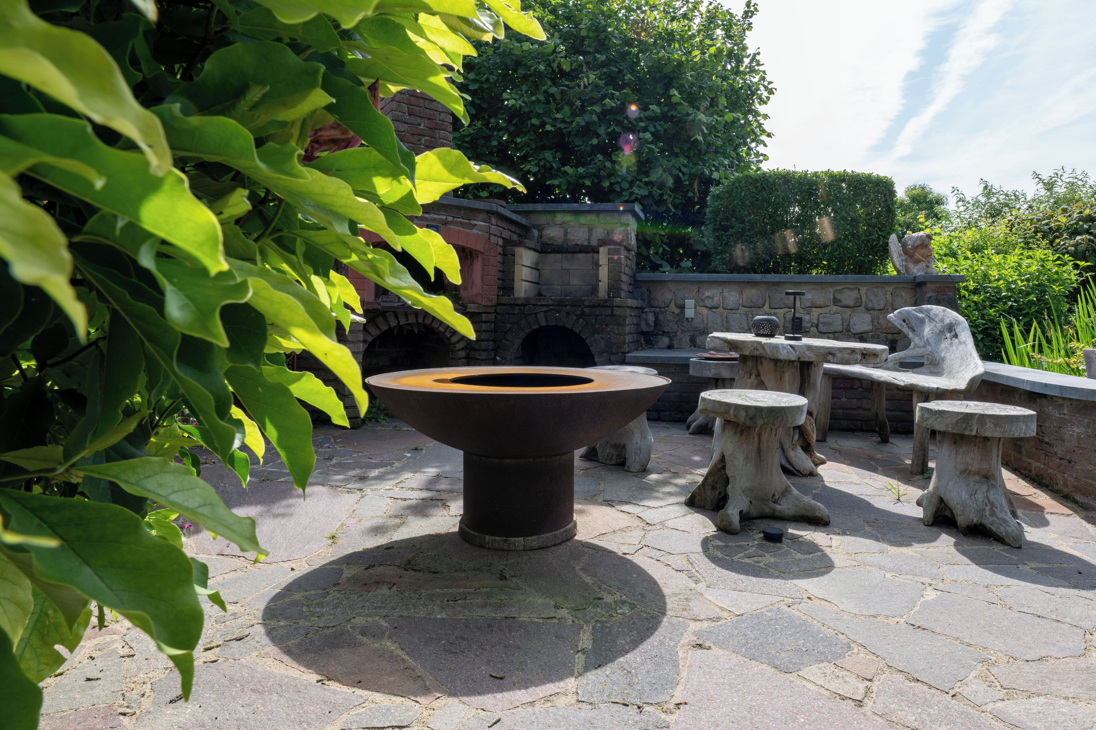 An outdoor patio area with stone flooring, a central fire pit, and rustic stone tables and benches.