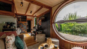 Un intérieur chaleureux avec une grande fenêtre ronde, des poutres en bois, un coin repas et un espace de vie comprenant un canapé et des coussins.
