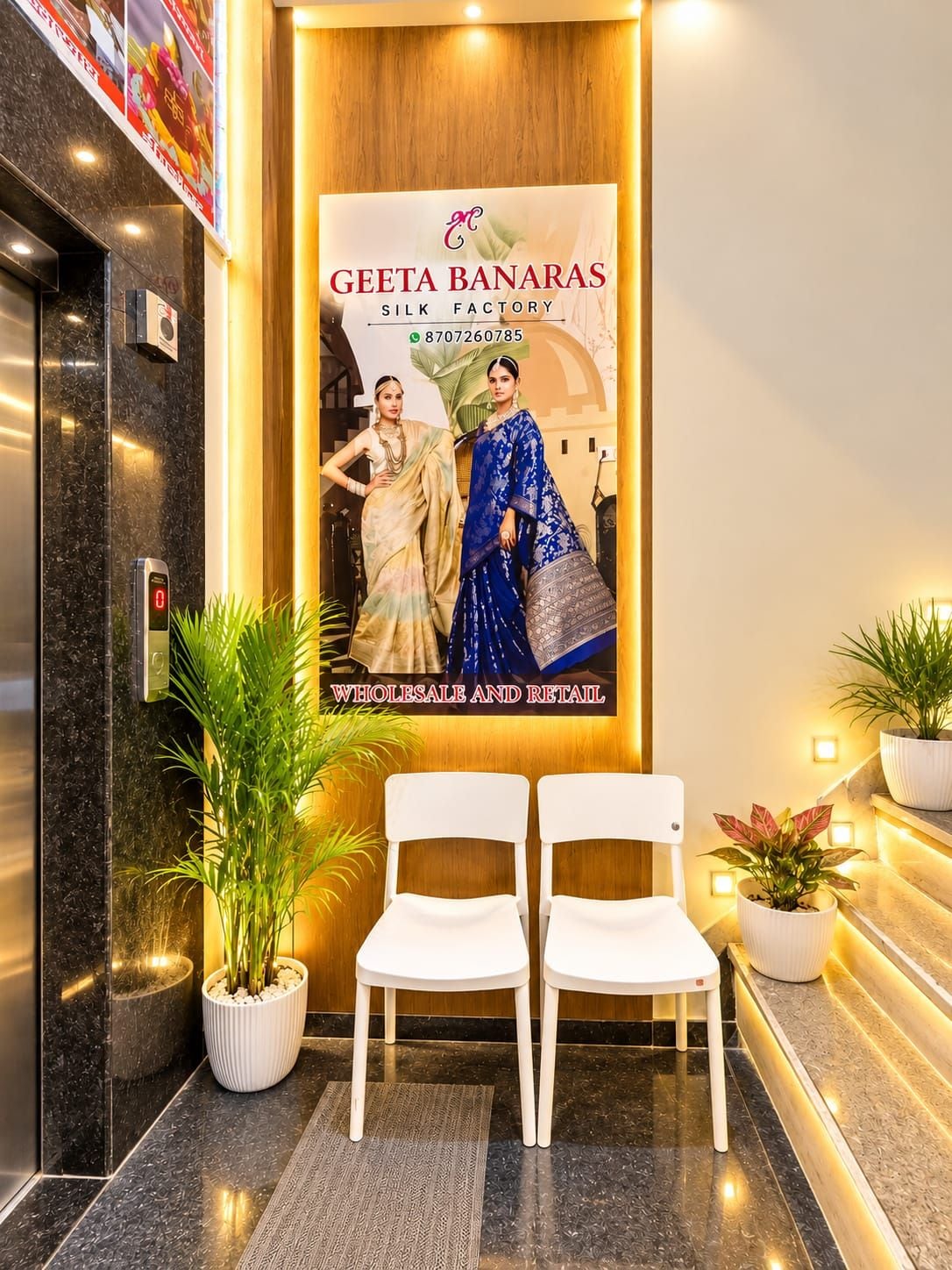 Un área de recepción con un cartel de Geeta Banaras Silk Factory, dos sillas blancas y plantas en macetas.