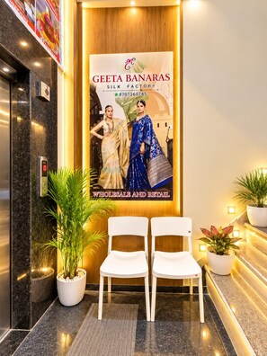 Un espace d’accueil avec une affiche de la Geeta Banaras Silk Factory, deux chaises blanches et des plantes en pot.