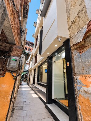 Un estrecho callejón con un edificio a un lado, que cuenta con fachada de vidrio y una puerta, y un muro de ladrillo al otro lado con tuberías y cables a la vista.