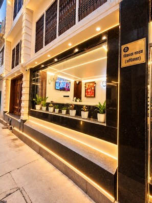 Entrada de un hotel con mostrador de recepción, plantas en macetas, un televisor y un cartel en idioma extranjero.