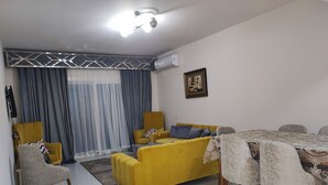 Un salon avec un canapé jaune, une table à manger recouverte de nappes en dentelle et un lustre décoratif au plafond.