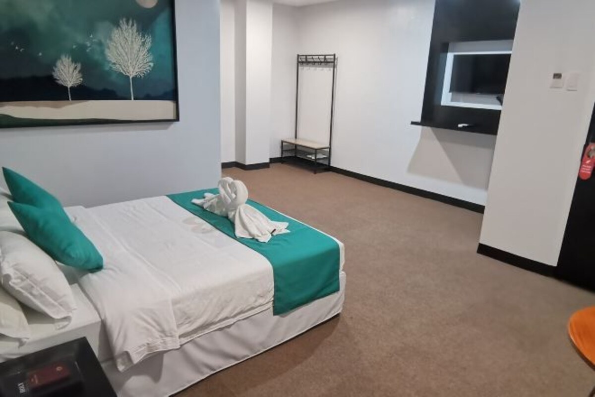 A hotel room with a bed, a TV, a coat rack, and a painting on the wall.