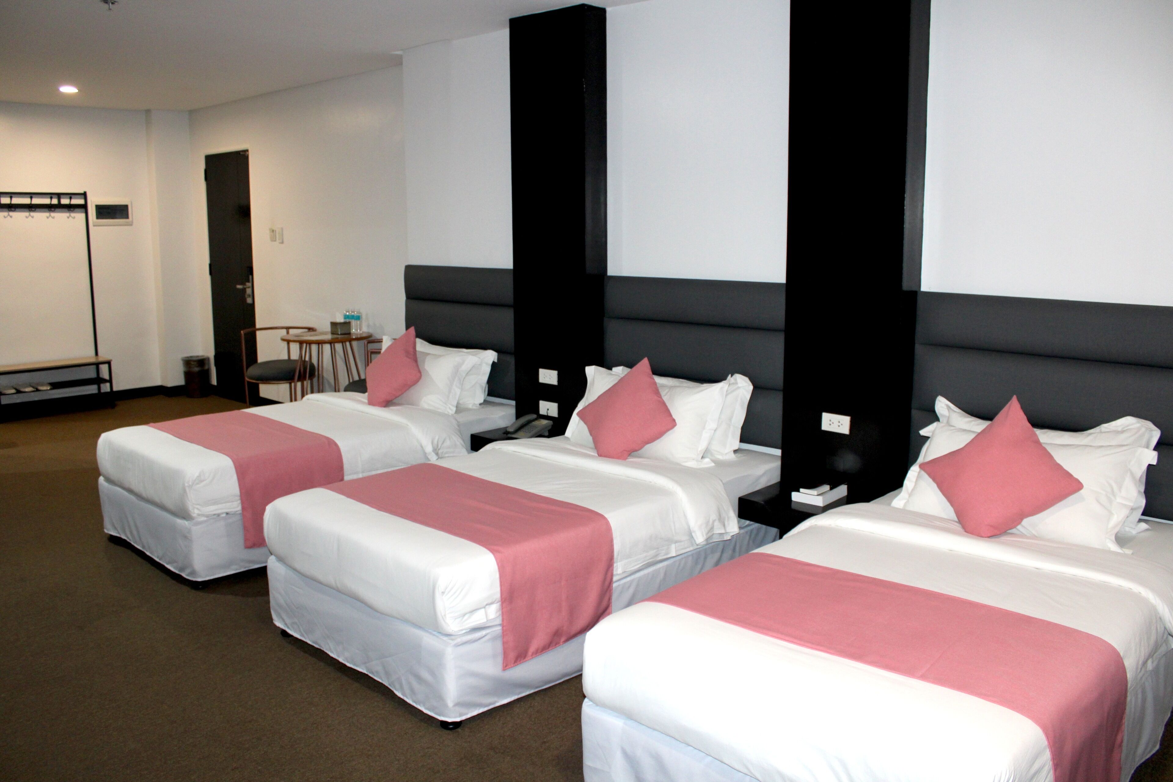 Ein Hotelzimmer mit drei Betten, jeweils mit weißer Bettwäsche und rosa Akzenten, einem dunklen Kopfteil und einem kleinen Tisch mit einem Stuhl.