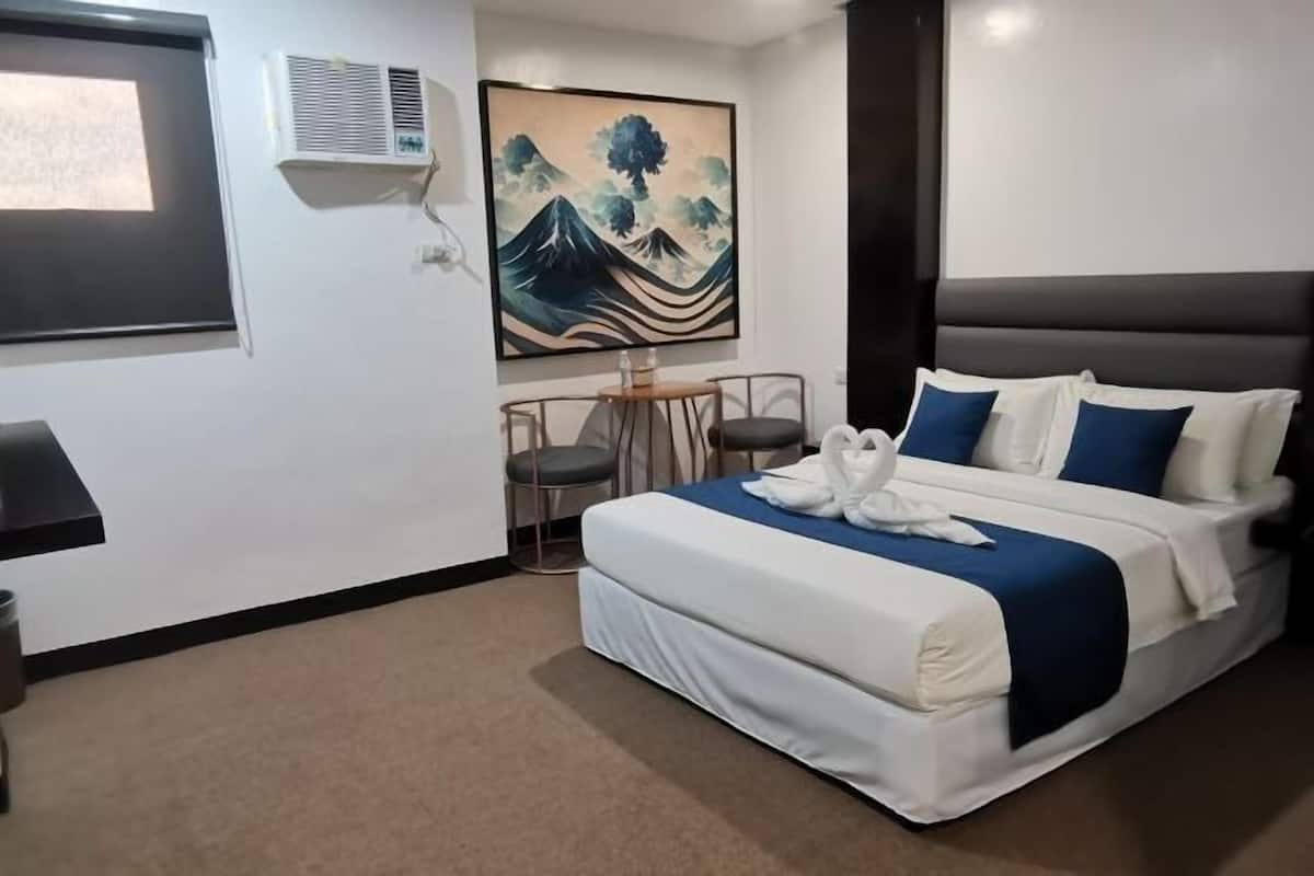 Una habitació d'hotel amb un llit, un quadre, una taula petita amb cadires i un aparell d'aire condicionat.