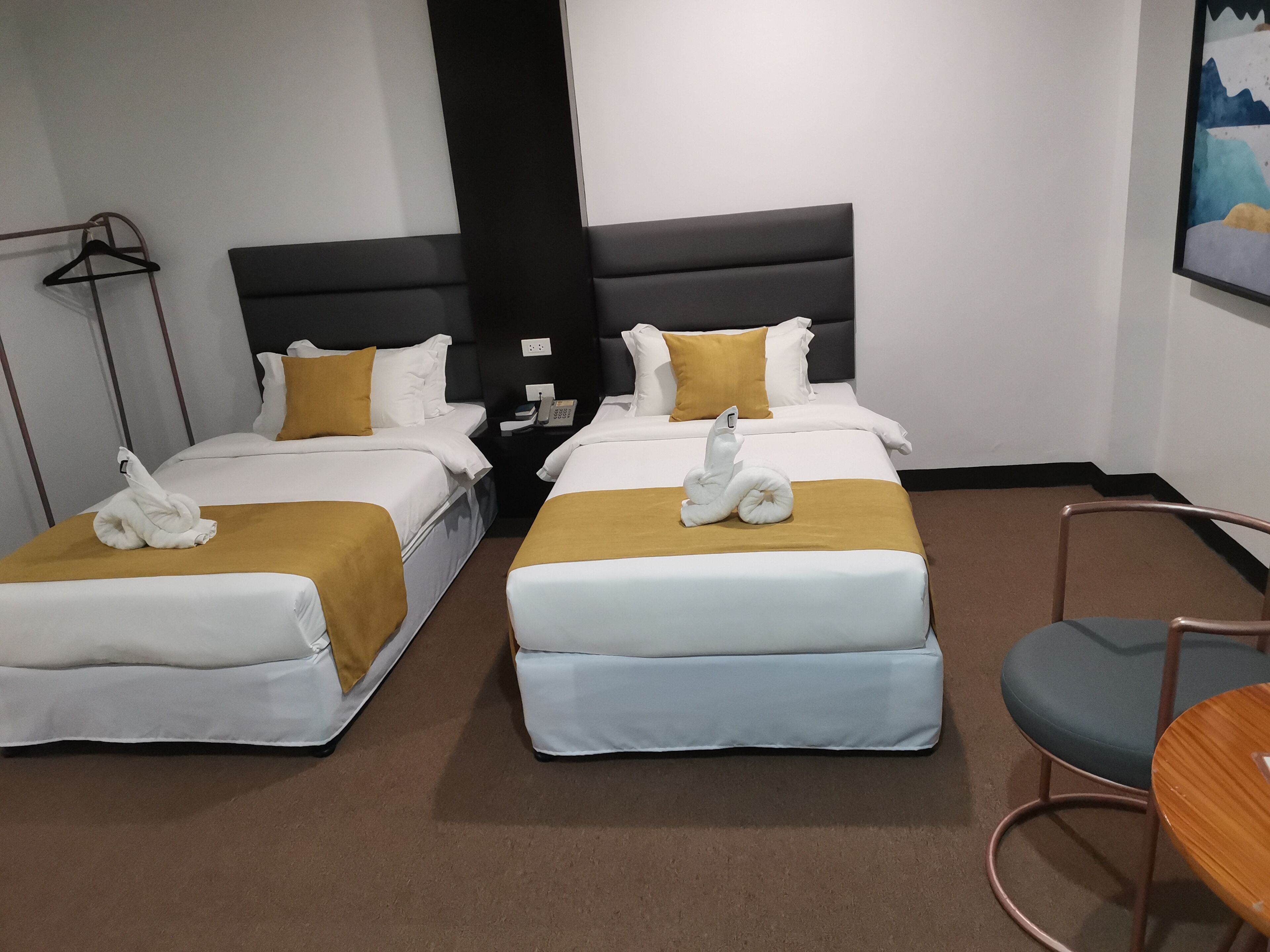 Habitación de hotel con dos camas, cada una con cubrecama blanco y dorado, y alfombra marrón. Hay una pequeña mesa con una silla, un teléfono instalado en la pared y un cuadro.