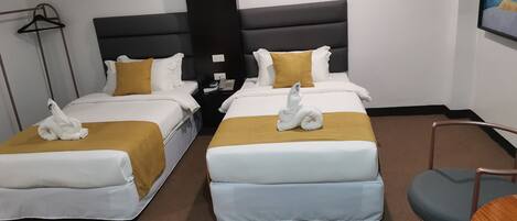 Habitación de hotel con dos camas, cada una con cubrecama blanco y dorado, y alfombra marrón. Hay una pequeña mesa con una silla, un teléfono instalado en la pared y un cuadro.