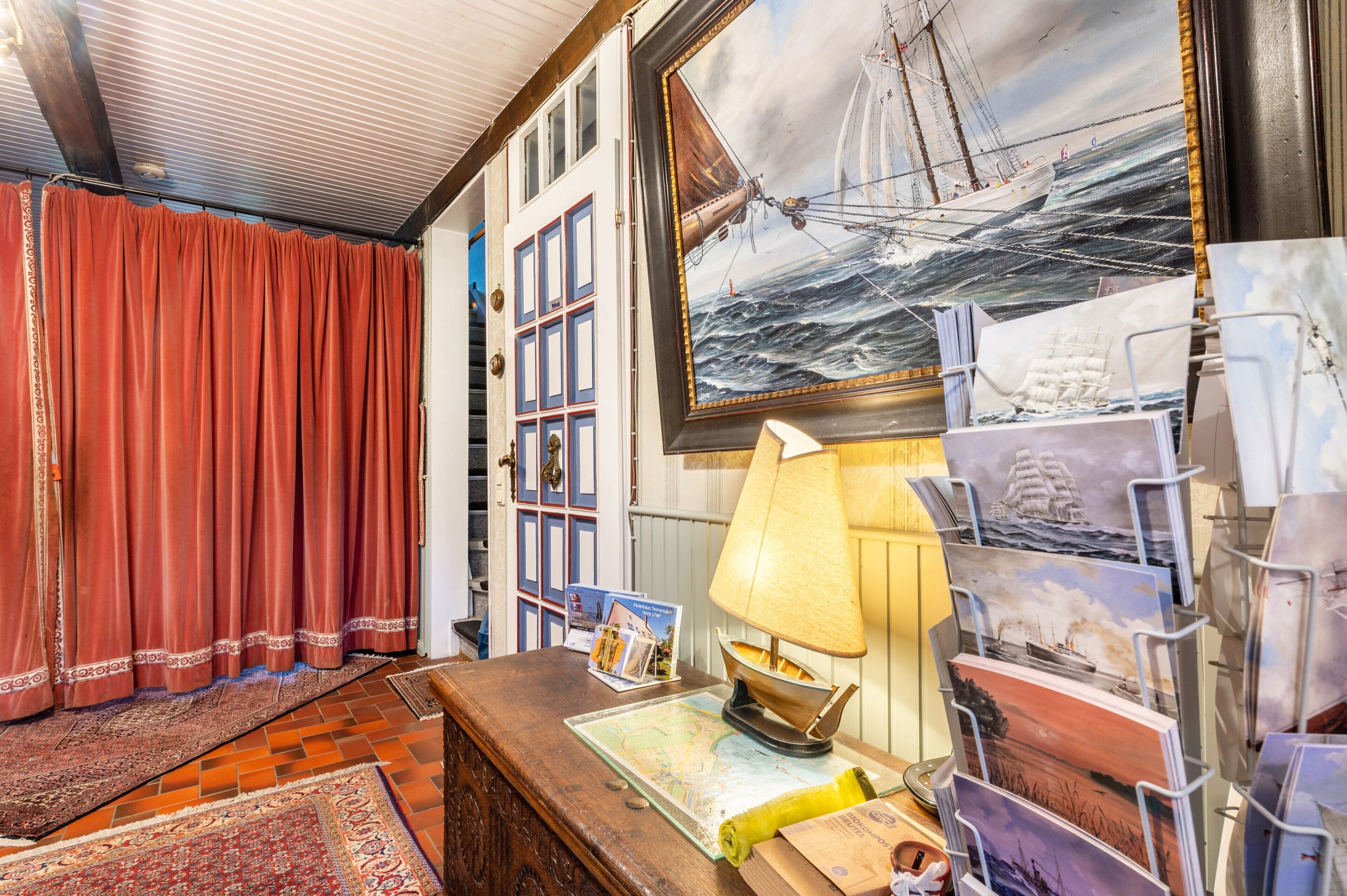 Une pièce dans laquelle on trouve un grand tableau représentant un navire sur la mer, un bureau en bois avec une lampe et un rideau rouge.