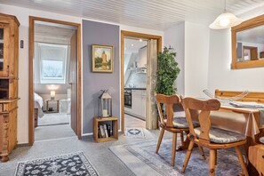 Una stanza con un tavolo da pranzo in legno, sedie, un piccolo tavolino con una lampada, un quadro appeso al muro e vista sulla cucina attraverso una porta aperta.