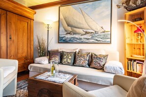 Una sala de estar con un mueble de madera, un sofá blanco con cojines estampados, una mesa de centro con una botella y un cuadro grande de un velero en la pared.