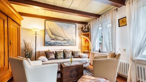 A cozy living room with a wooden cabinet, a sofa with patterned cushions, a coffee table with a decorative box, and a large sailboat painting on the wall. - Apartment 'Scharhörn' with Shared Terrace, Shared Garden and Wi-Fi (Cuxhaven)