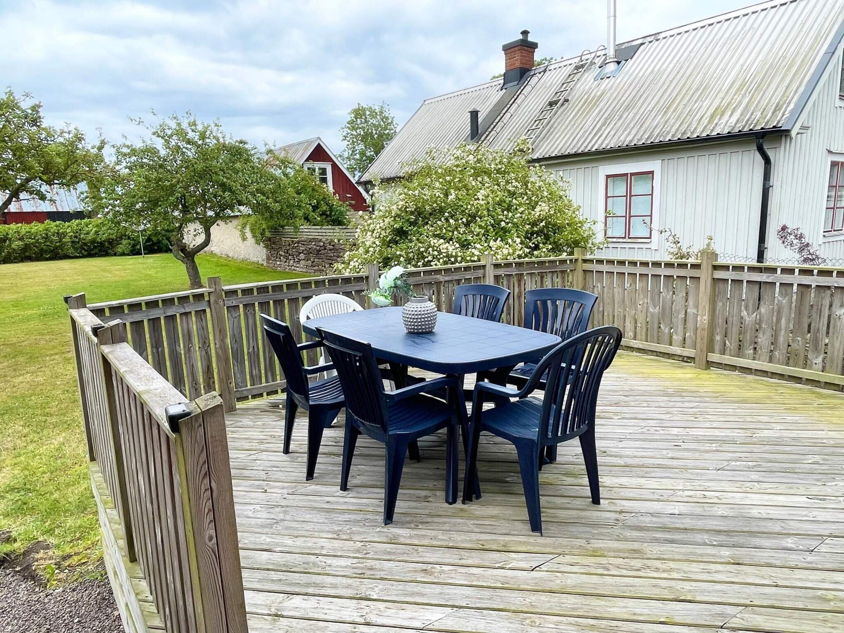 Una terraza de madera con mesa y sillas, rodeada por una valla de madera, y una casa con techo de metal al fondo.