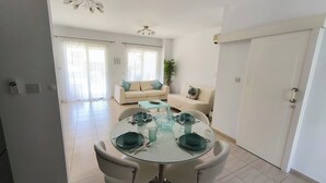 A dining area with a round glass table set for four, surrounded by white chairs. There is a sofa and a small table in the background. - Sophia Bay Home - a treasure box in Peyia (Pegeia)