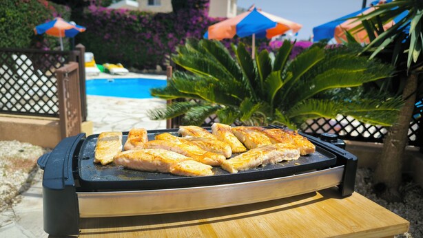 A grill with several pieces of grilled chicken in an outdoor setting with a pool and umbrellas in the background. - Sophia Bay Home - a treasure box in Peyia (Pegeia)