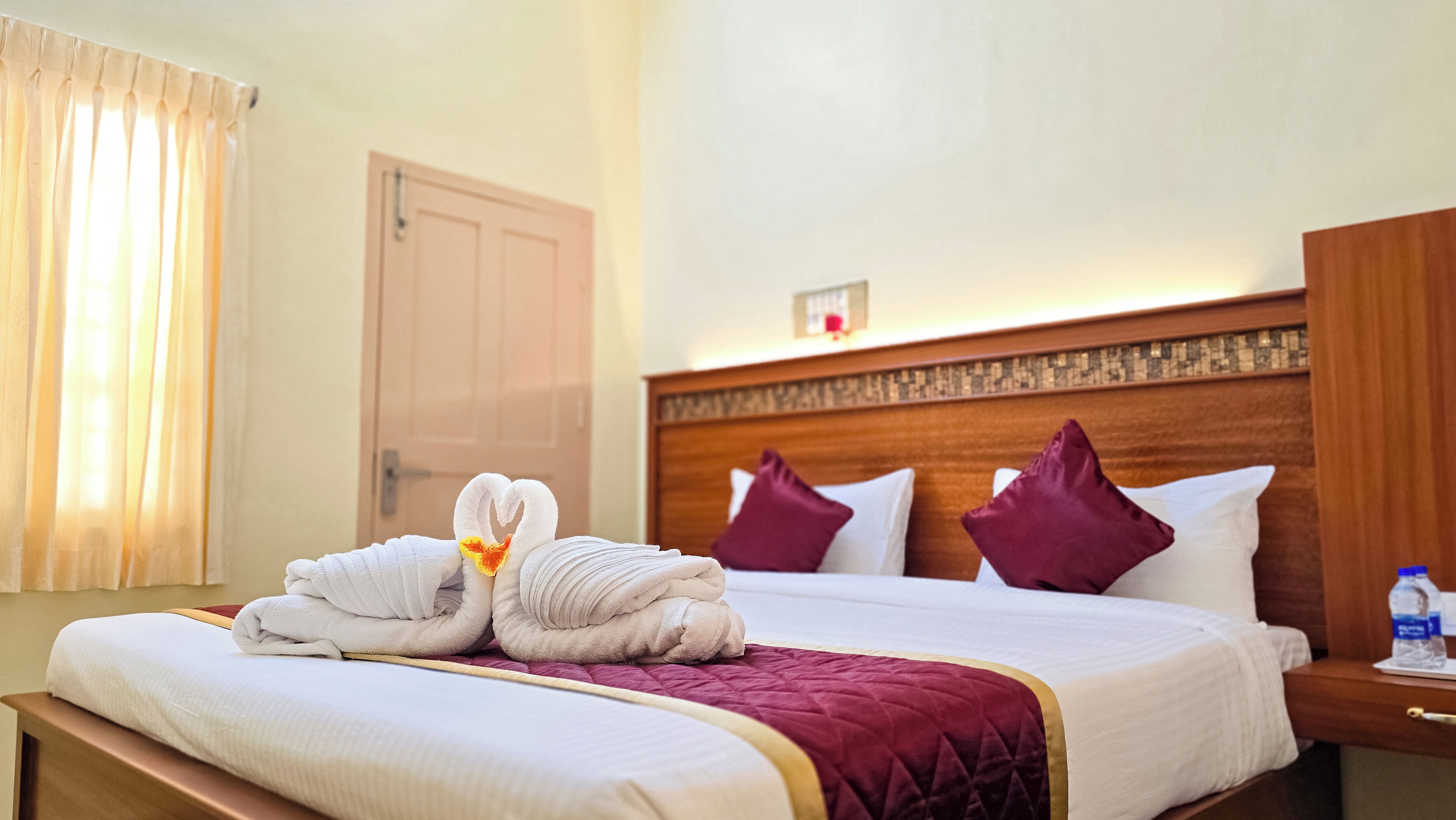 A hotel room with a neatly made bed, a wooden headboard, and a nightstand with a bottle and glass.