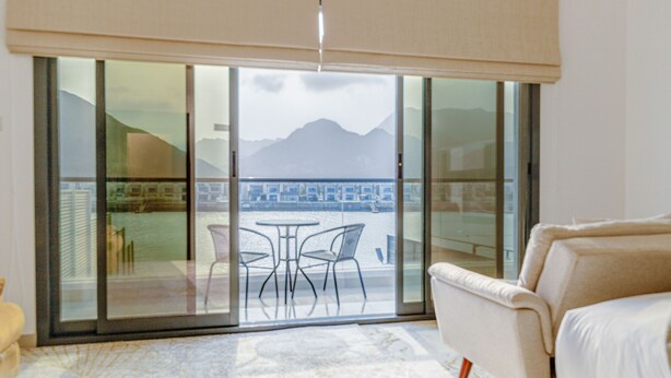 A room with a sliding glass door leading to a balcony with a view of a cityscape and mountains. There is a white sofa and a small table with chairs. - Villa with Jacuzzi and Beach Access, Near Diving, Paddle Boating and Camping Spots by Deluxe Holiday Homes (Fujairah)
