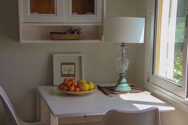 A room with a white table, a bowl of fruit, a lamp, and a window with a view of greenery.