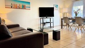 A modern living room with a glass coffee table, a flat-screen TV, a sofa, and a dining area with chairs and a table.