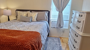 Une chambre à coucher avec un lit, une commode, une table de chevet, une lampe et une fenêtre avec des rideaux.
