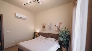A bedroom with a bed, an air conditioner, a nightstand with a lamp, and wall art. - Hazel Spaces In New Cairo (New Cairo)