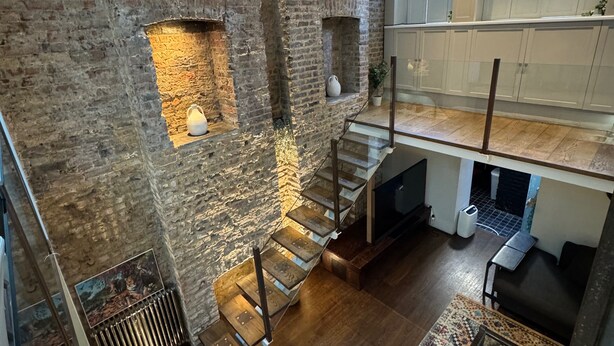 A loft-style room with exposed brick walls, a wooden staircase, and a mezzanine level with white cabinets.