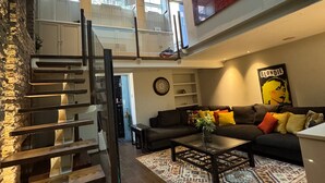 A modern loft apartment with a staircase, a living area with a sofa and coffee table, and a large window providing natural light.