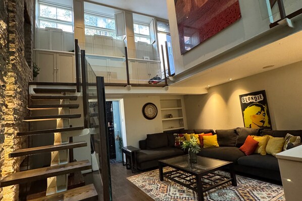 A modern loft apartment with a staircase, a living area with a sofa and coffee table, and a large window providing natural light. - Covent Garden House, 4-bed, Sleeps 10 (London)