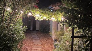 A wooden pathway leading through a garden at night, surrounded by lush greenery and illuminated by string lights.