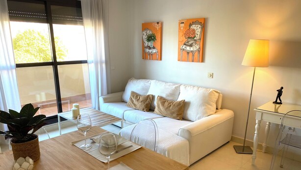 Un salon moderne avec un canapé blanc, une table à manger dressée avec des assiettes, des verres et des couverts, et une chaise transparente. Deux tableaux ornent le mur, une lampe est posée sur une table d’appoint, et une grande fenêtre est agrémentée de rideaux.