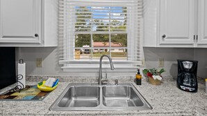 A kitchen with a double sink, white cabinets, a window with blinds, and a coffee maker.