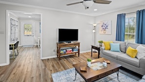 A living room with a ceiling fan, a flat-screen TV, a wooden coffee table, a gray sofa with yellow and blue cushions, and a dining area with a white table and chairs.