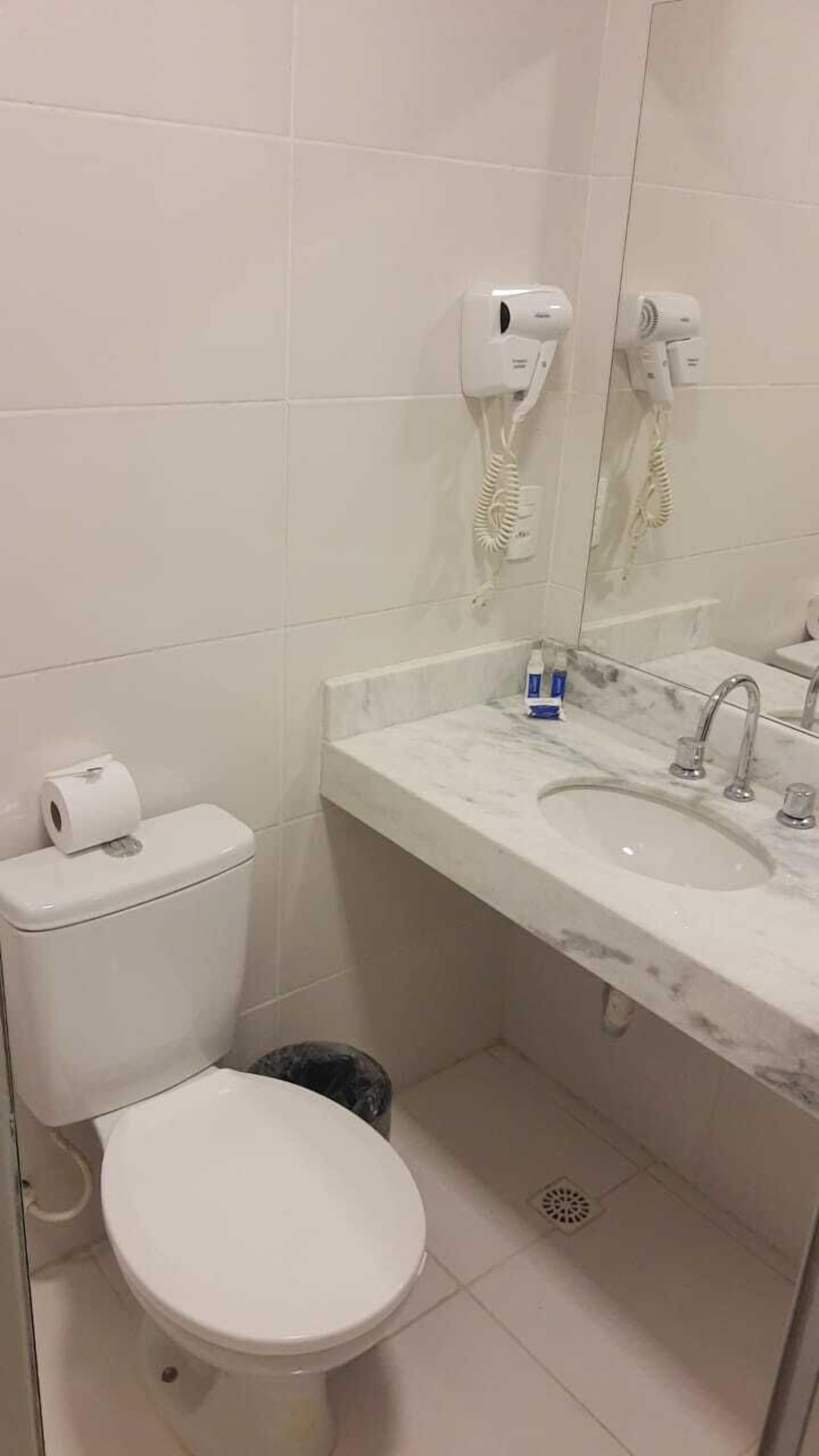 Un baño con inodoro, lavamanos y secador de pelo montado en la pared.