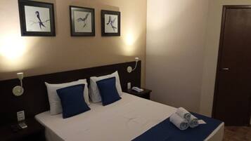 Quarto de hotel com uma cama, duas almofadas, uma mesinha de cabeceira, um telefone, um quadro na parede e uma porta.