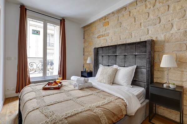 A bedroom with a stone wall, a bed with a headboard, a nightstand, a lamp, and a window with curtains.