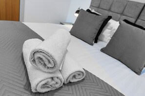 Un letto rifatto con cura, asciugamani bianchi arrotolati, biancheria da letto grigia fantasia e cuscini grigi.