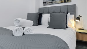 Una cama bien hecha con toallas enrolladas, ropa de cama gris y un cuadro en la pared.