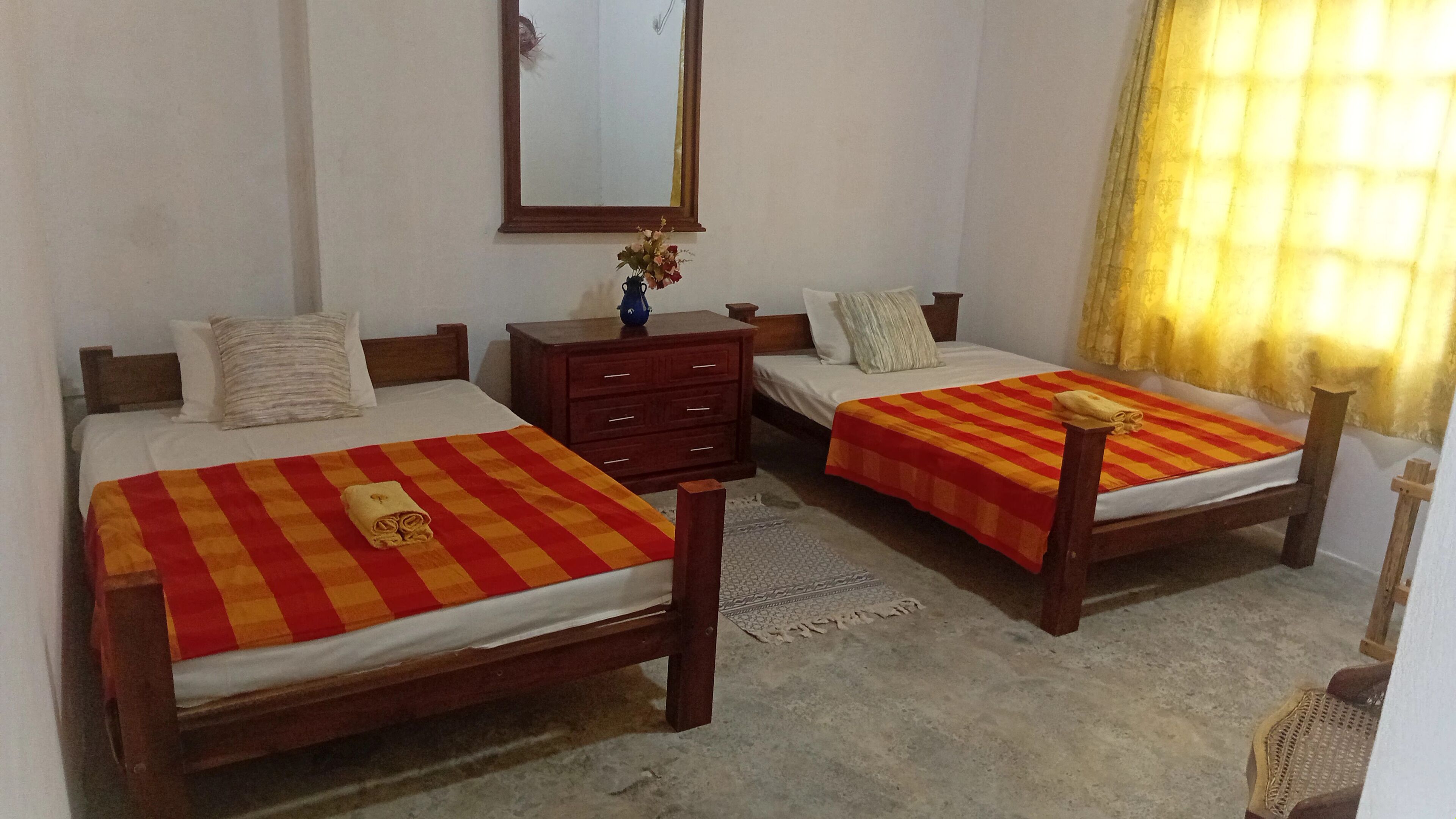A hotel room with two wooden beds, each with a red and yellow striped bedspread and a rolled towel on one side. There is a wooden nightstand with a vase of flowers between the beds.