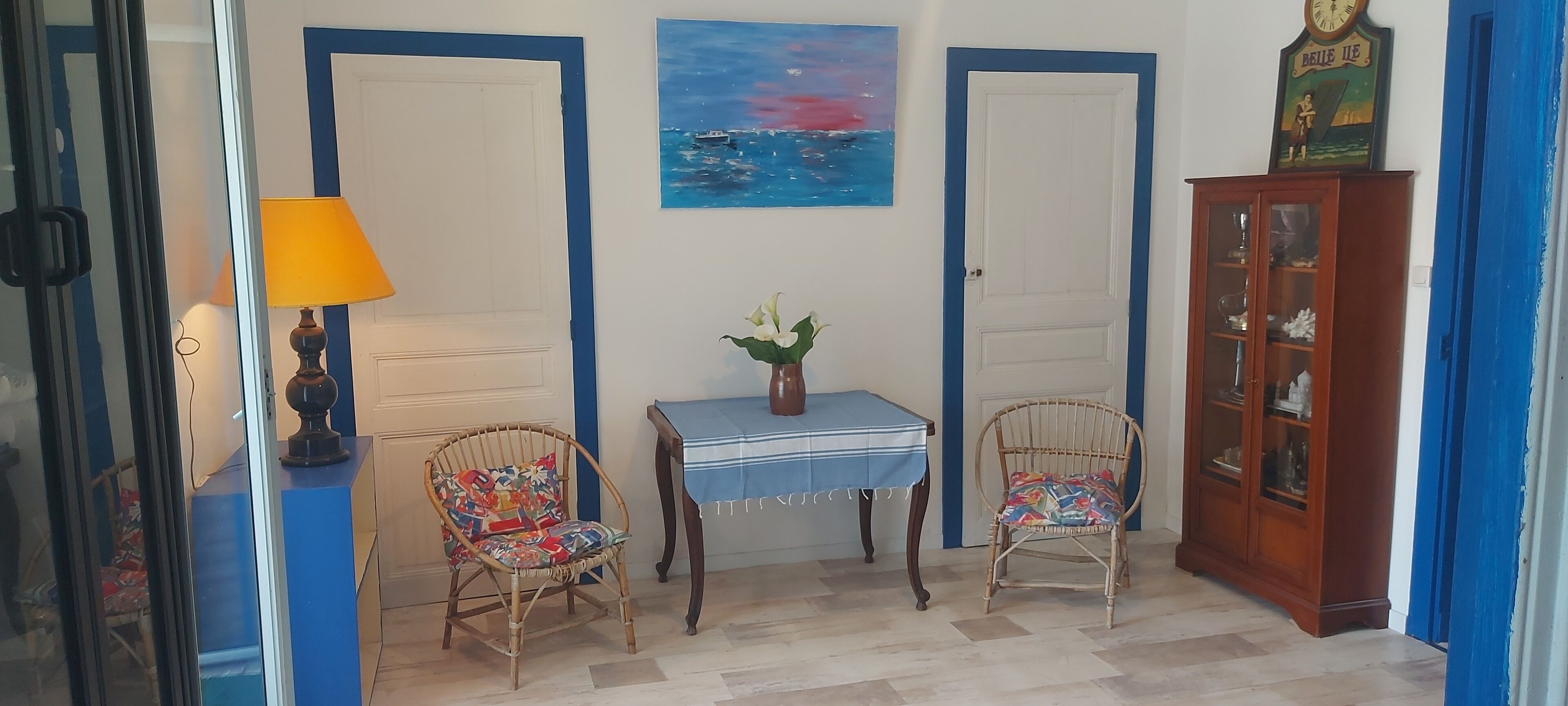 Una habitación con dos puertas, una mesa con un florero, dos sillas de mimbre, una lámpara y un mueble con puertas de vidrio.
