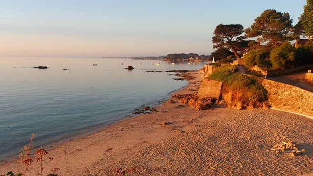 A coastal beach with calm waters, boats, and a stone wall with vegetation.