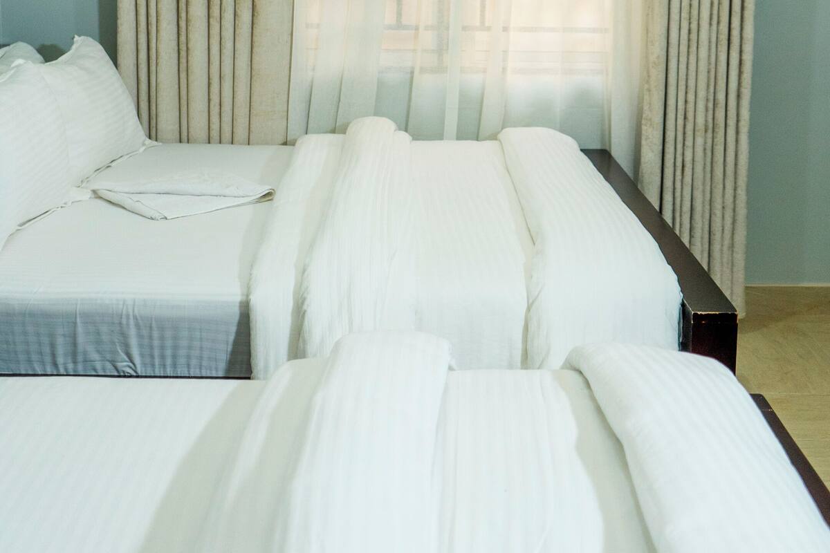Ein Hotelzimmer mit zwei Betten, einem Fenster mit durchsichtigen Vorhängen und einem Holzkopfende.