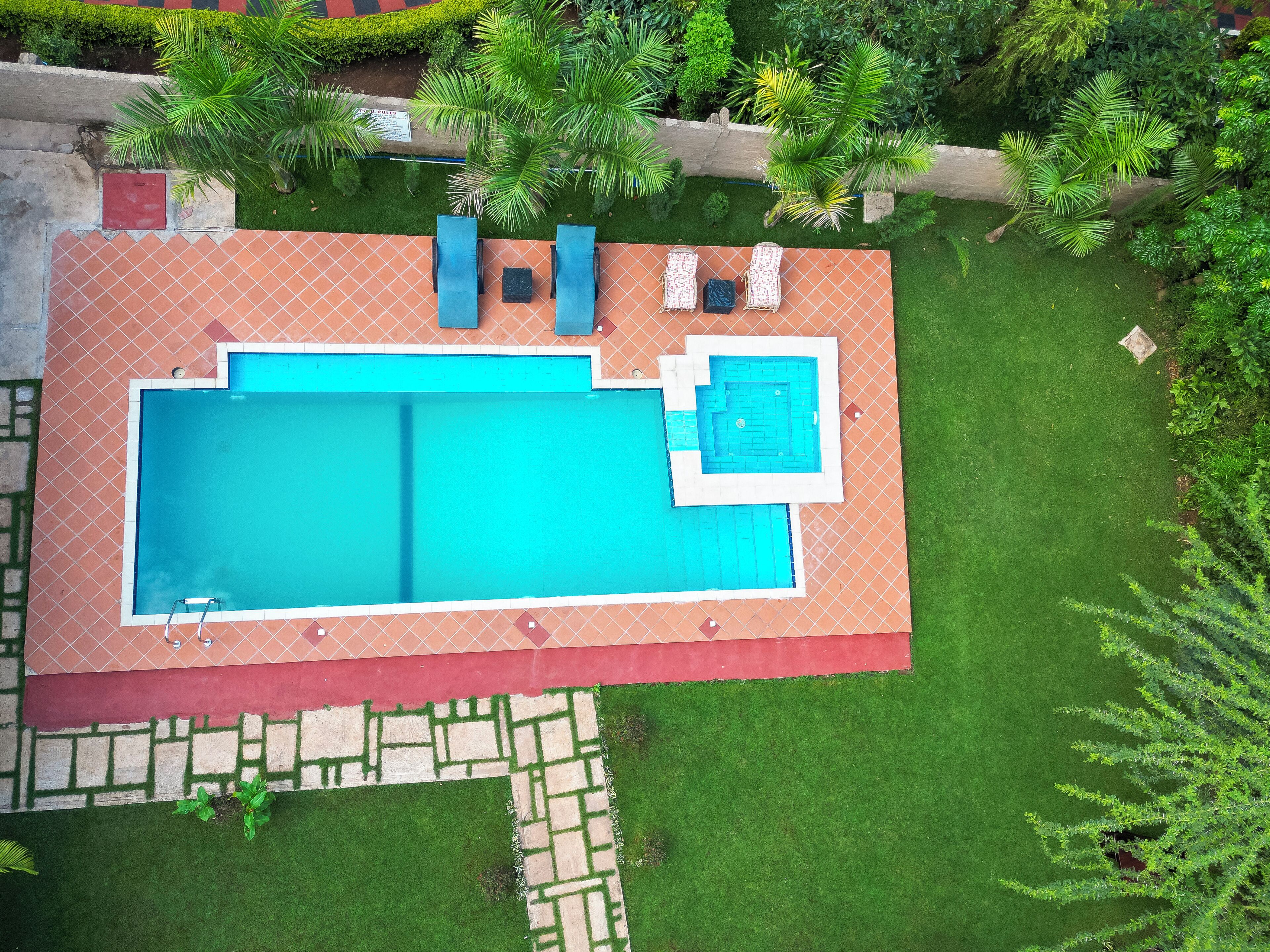 Ein rechteckiger Pool mit einem kleineren angrenzenden Pool, umgeben von einer gefliesten Terrasse und einer Rasenfläche mit einem Weg.