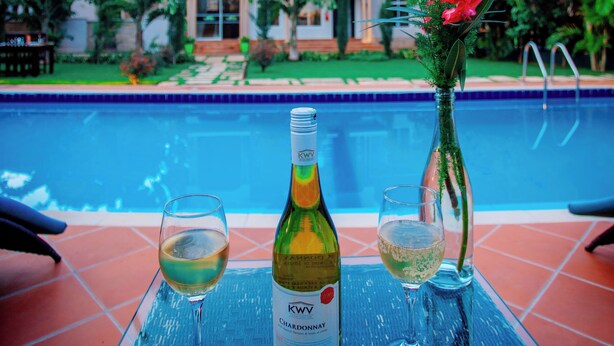 Mesa à beira da piscina com uma garrafa de vinho e dois copos, uma casa ao fundo e céu claro.