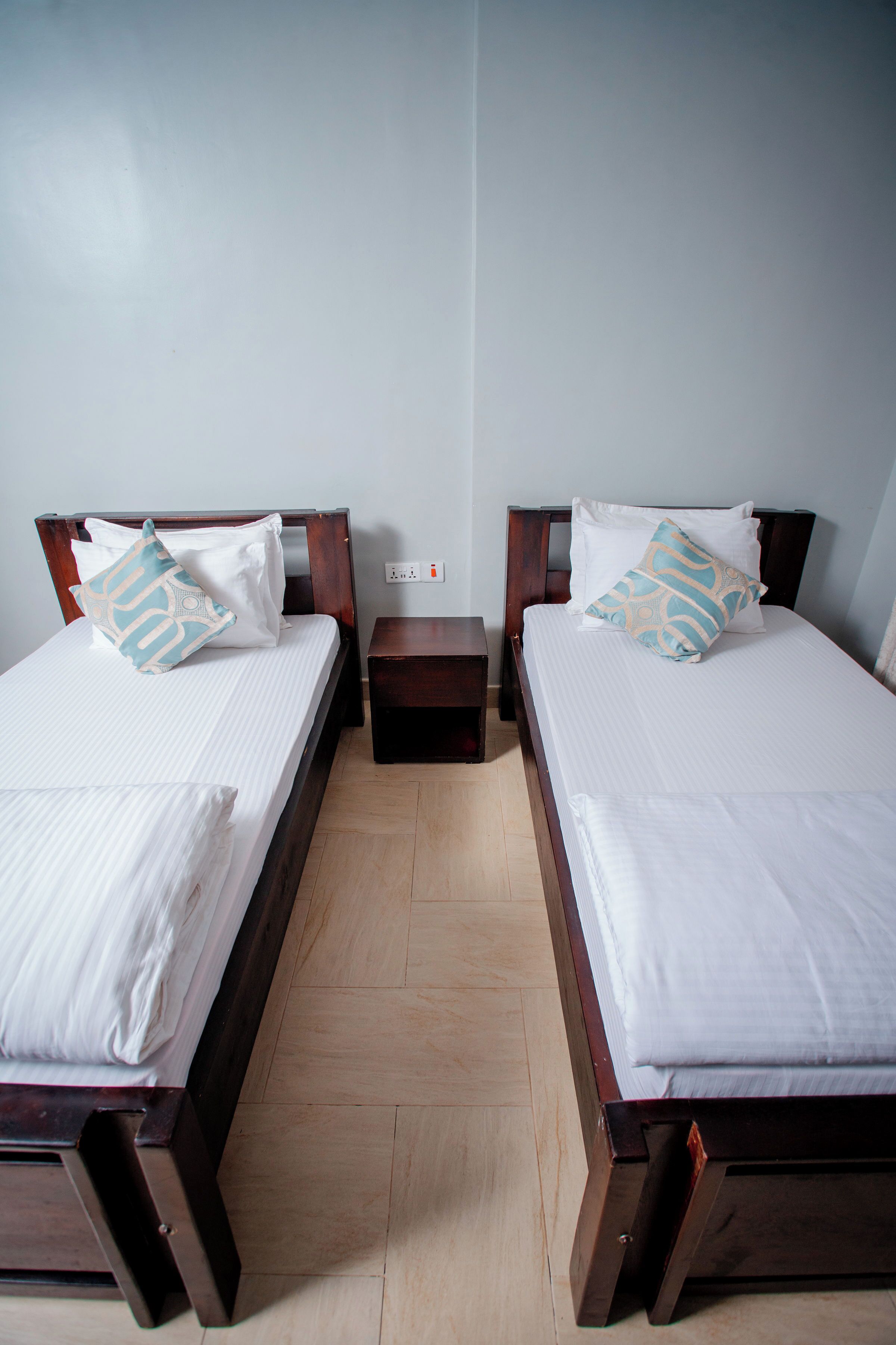 A hotel room with two single beds, wooden headboards, white linens, and a small bedside table.