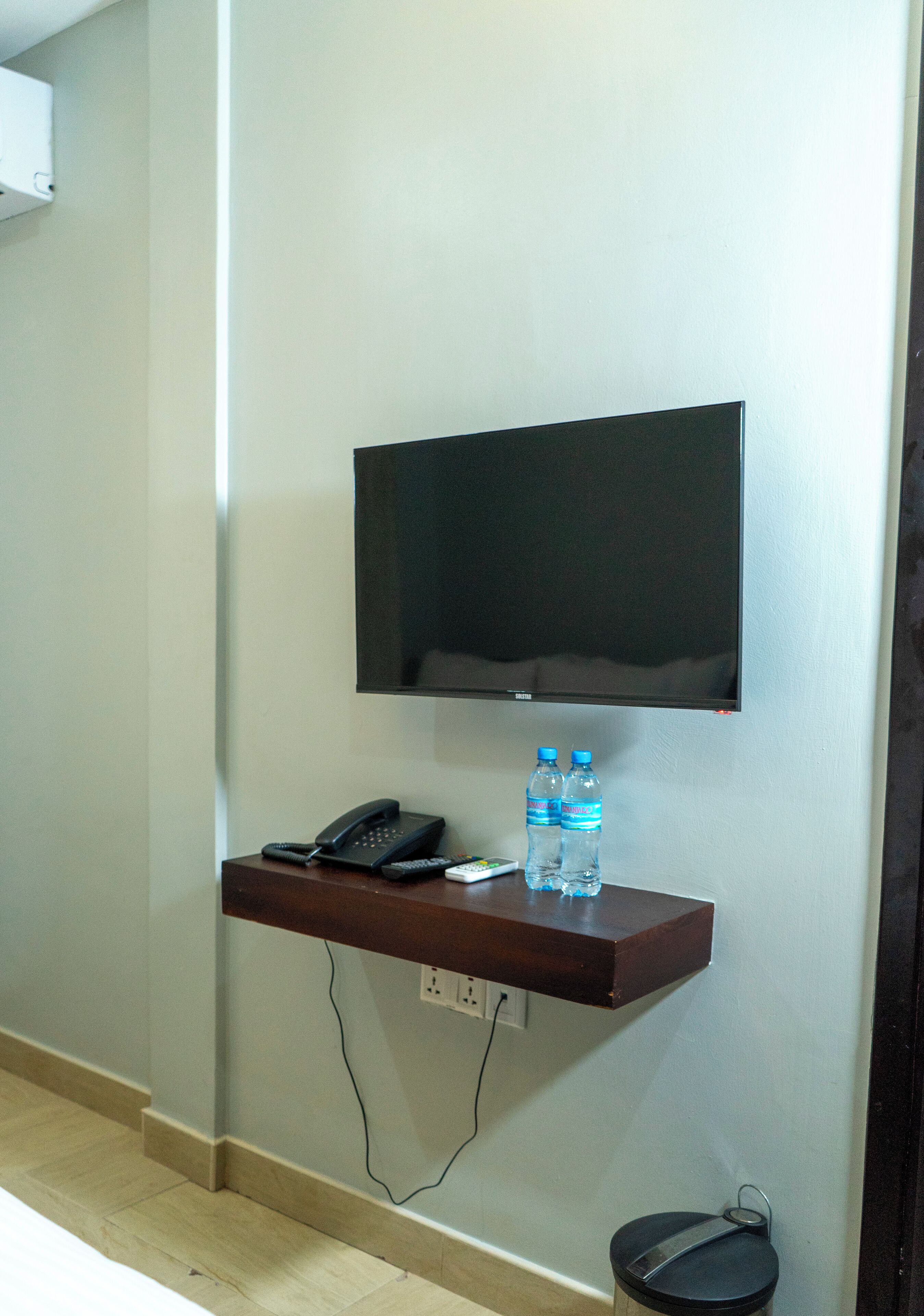 Una TV a parete sopra un ripiano in legno con un telefono, telecomandi e due bottiglie d'acqua.